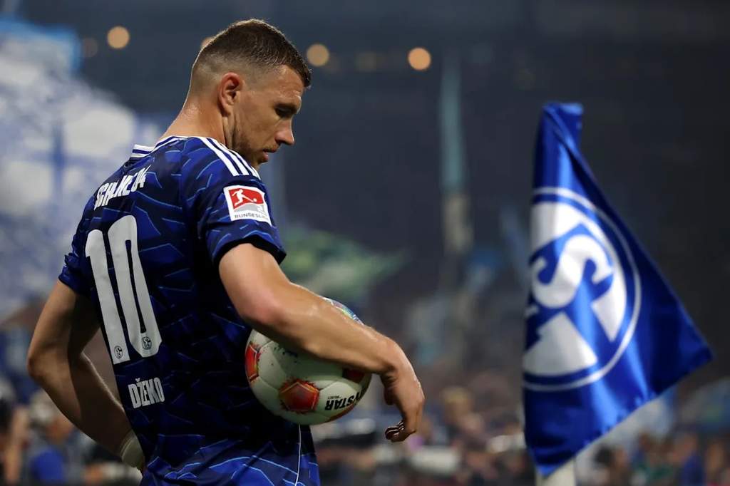 Penyerang berpengalaman asal Bosnia, Edin Dzeko, mengenakan perlengkapan latihan klub sepak bola dengan latar belakang stadion megah.