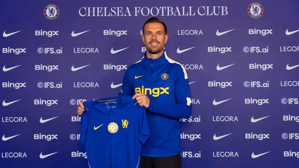 Jordan Henderson mengenakan jersey latihan Chelsea di kompleks latihan Stamford Bridge dengan latar lapangan hijau.
